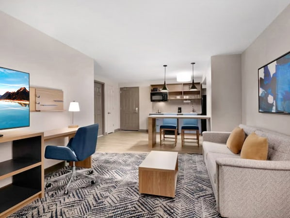 Une chambre d’hôtel moderne équipée d’un téléviseur à écran plat, d’un bureau avec une chaise, d’un canapé, d’une table basse, d’un tapis à motifs et d’une kitchenette à l’arrière-plan.