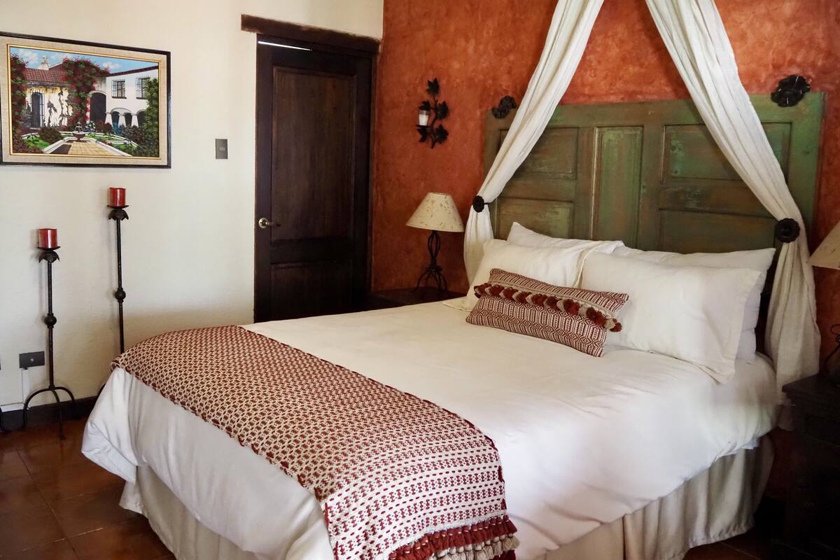 A bedroom with a four-poster bed, a painting on the wall, and a door.