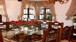 Un comedor con mesa de vidrio rodeada de sillas de madera, ventanas amplias con cortinas y plantas en macetas.