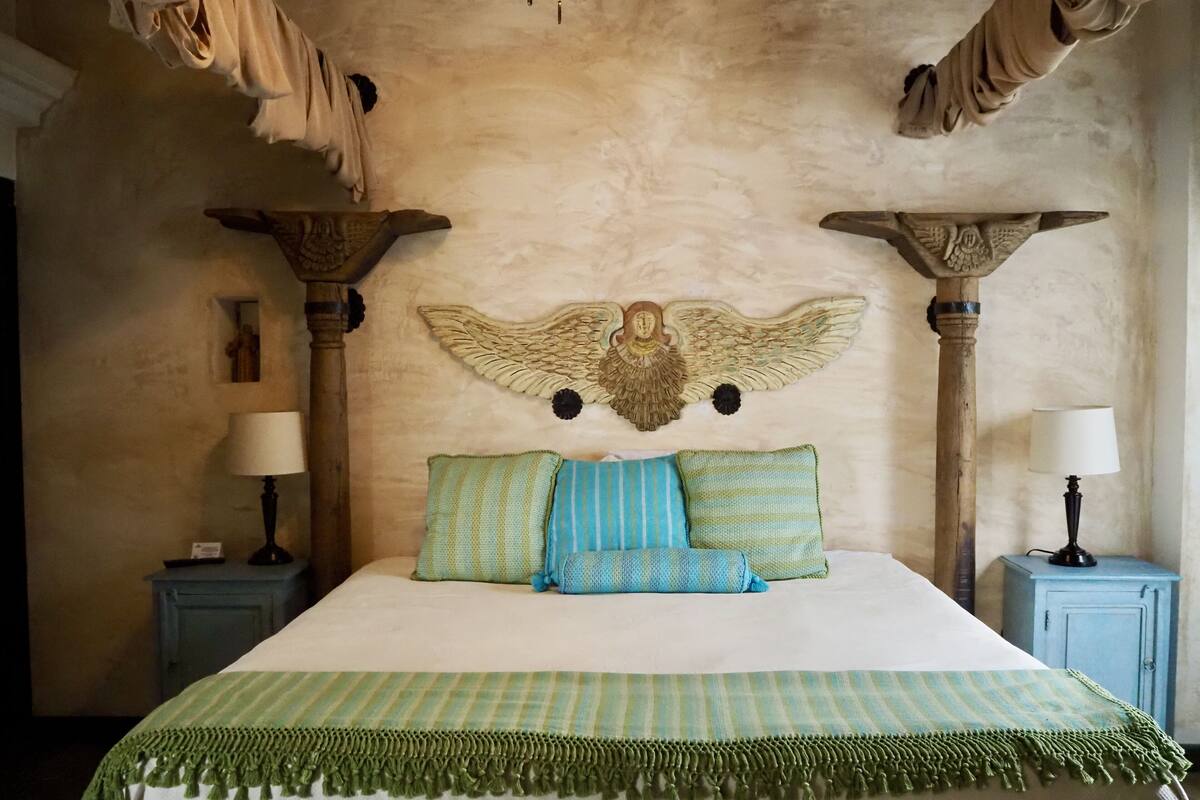 A bed with a green and white striped bedspread, two pillows, and a green fringe. There are two nightstands with lamps, and a wall with a decorative angel headboard.