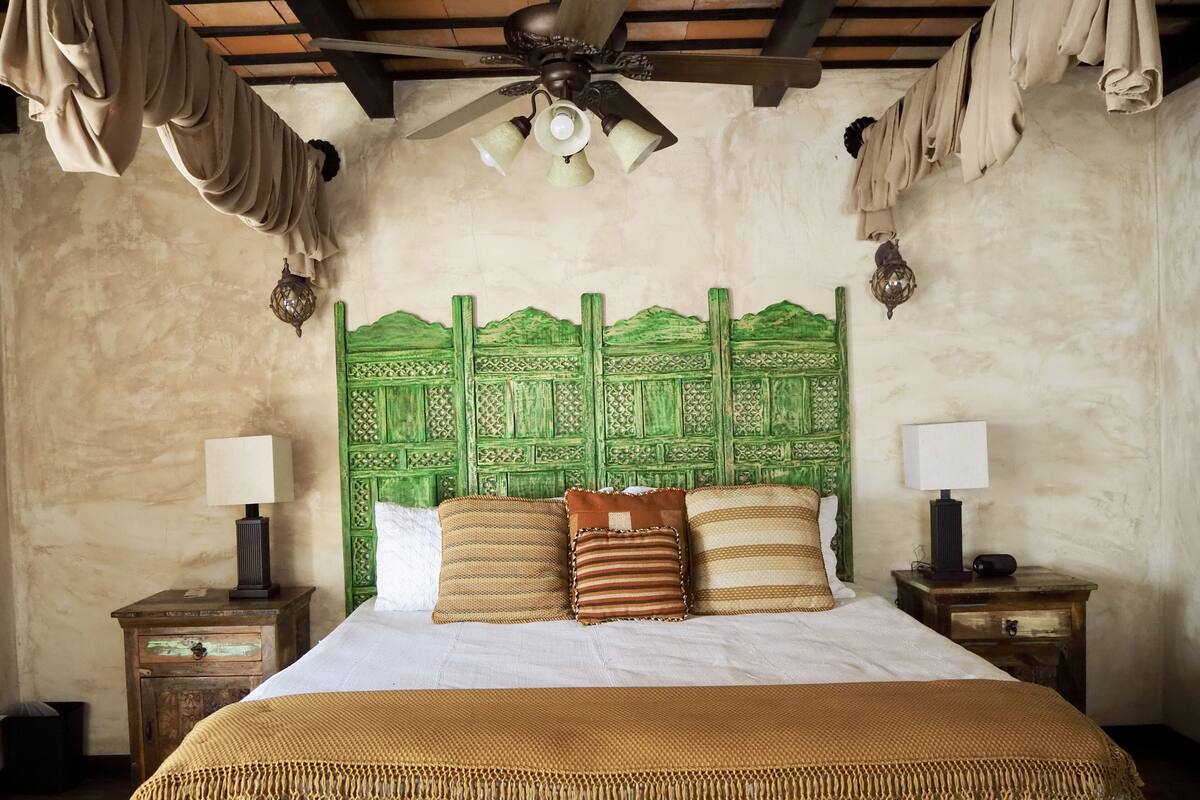 A bedroom with a green headboard, two bedside tables with lamps, a bed with a white and brown blanket, and a ceiling fan.