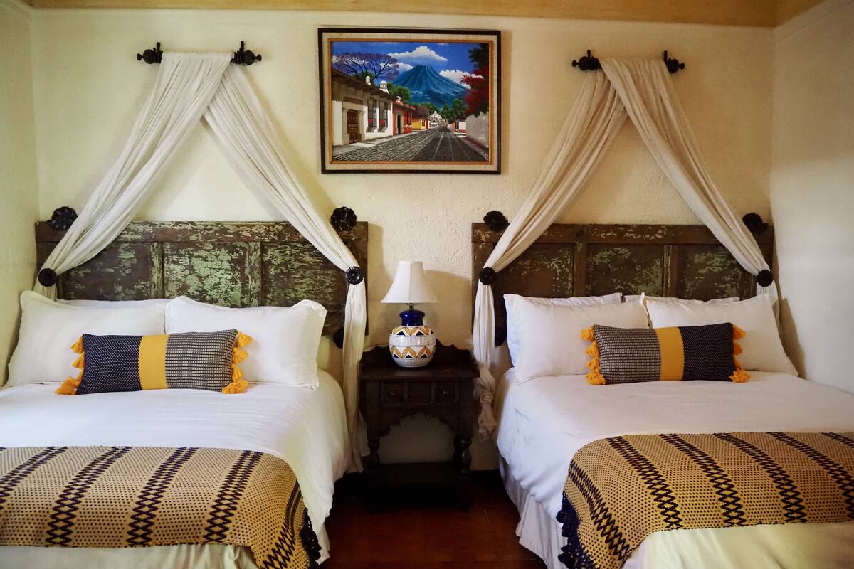 A room with two beds, each with a canopy, a nightstand with a lamp, and a framed painting on the wall.