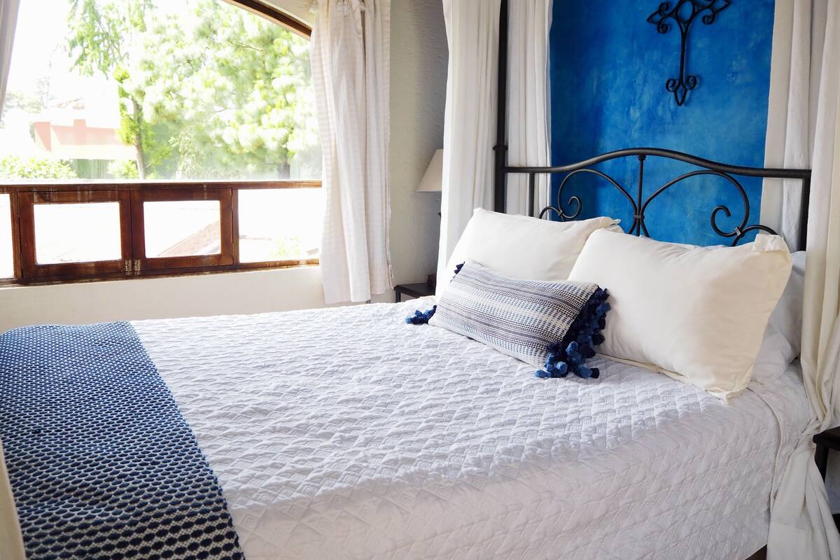 A bed with white bedding and a blue patterned throw, a blue wall with a cross decoration, and a window with curtains.