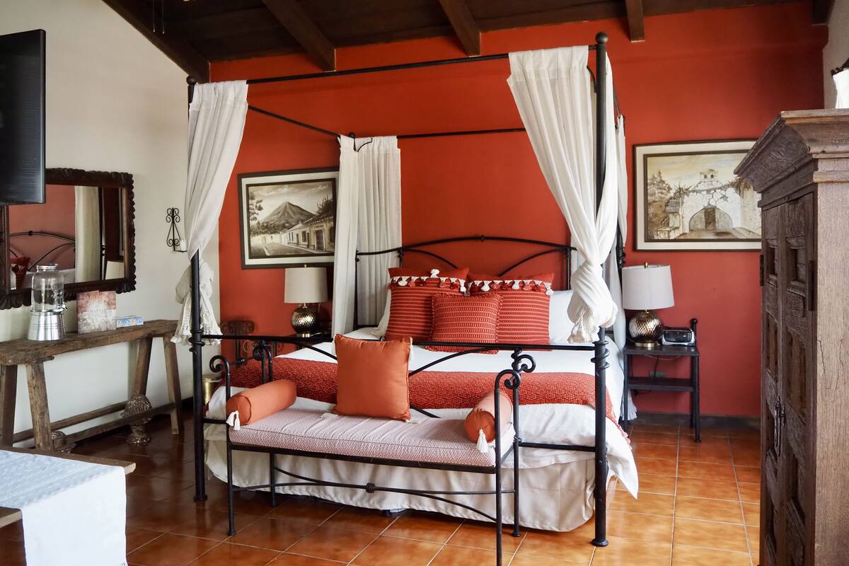 A four-poster canopy bed with a red and white bedspread, two nightstands with lamps, a wooden nightstand with a glass container, a mirror, and two framed pictures on the wall.