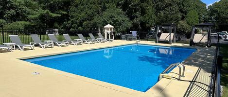 Una piscina rectangular con superficie de agua azul cristalina, rodeada por una zona de cubierta color beige y sombras de árboles.