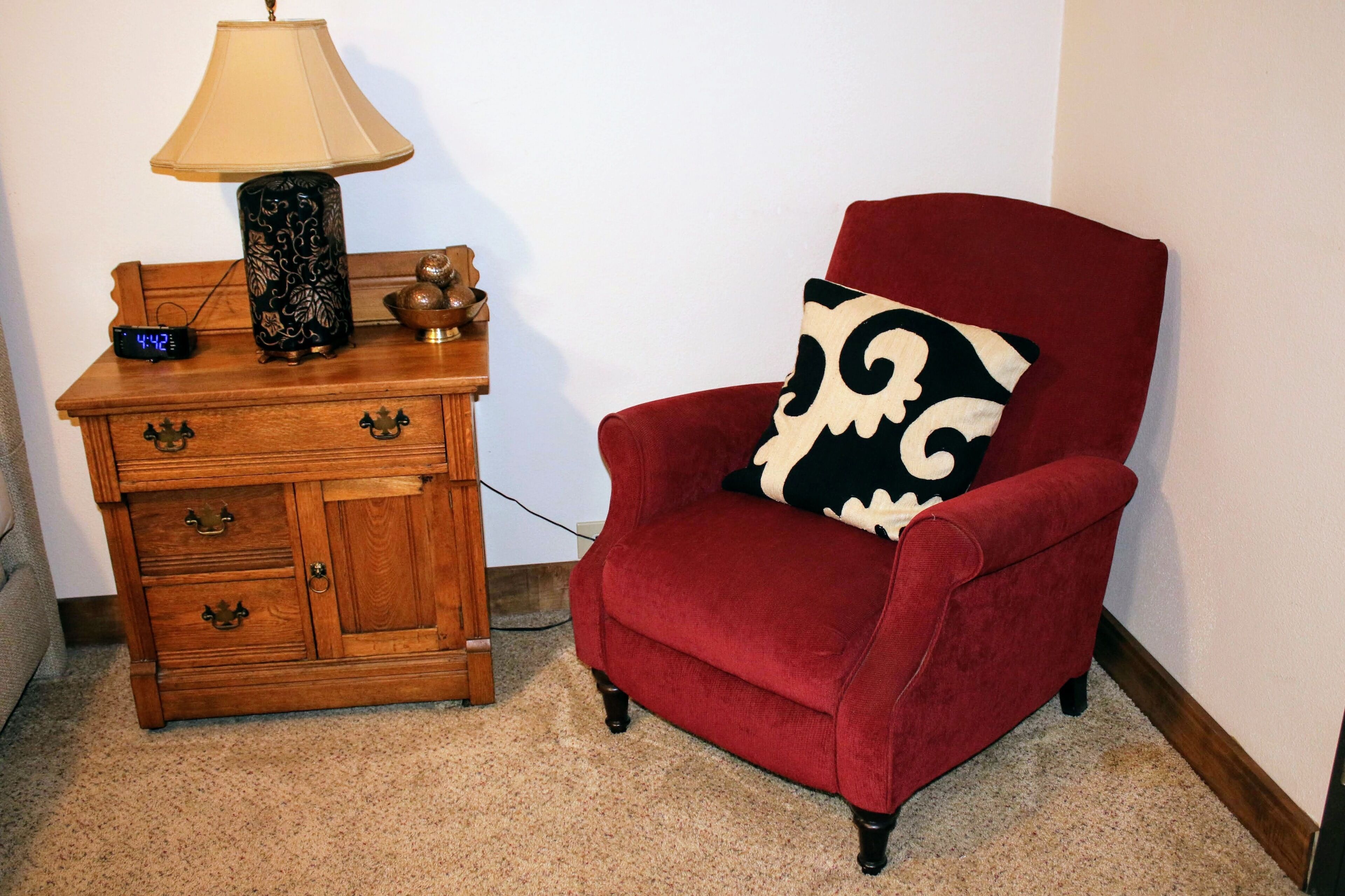 A red armchair with a patterned pillow, a wooden side table with a lamp, and a digital clock.