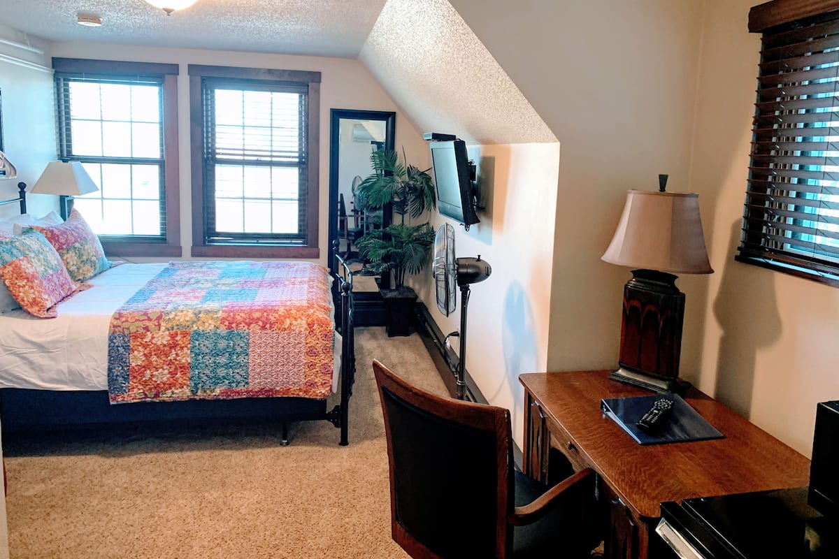 Un dormitorio con cama, escritorio, silla, lámpara, televisor y ventana con persianas.