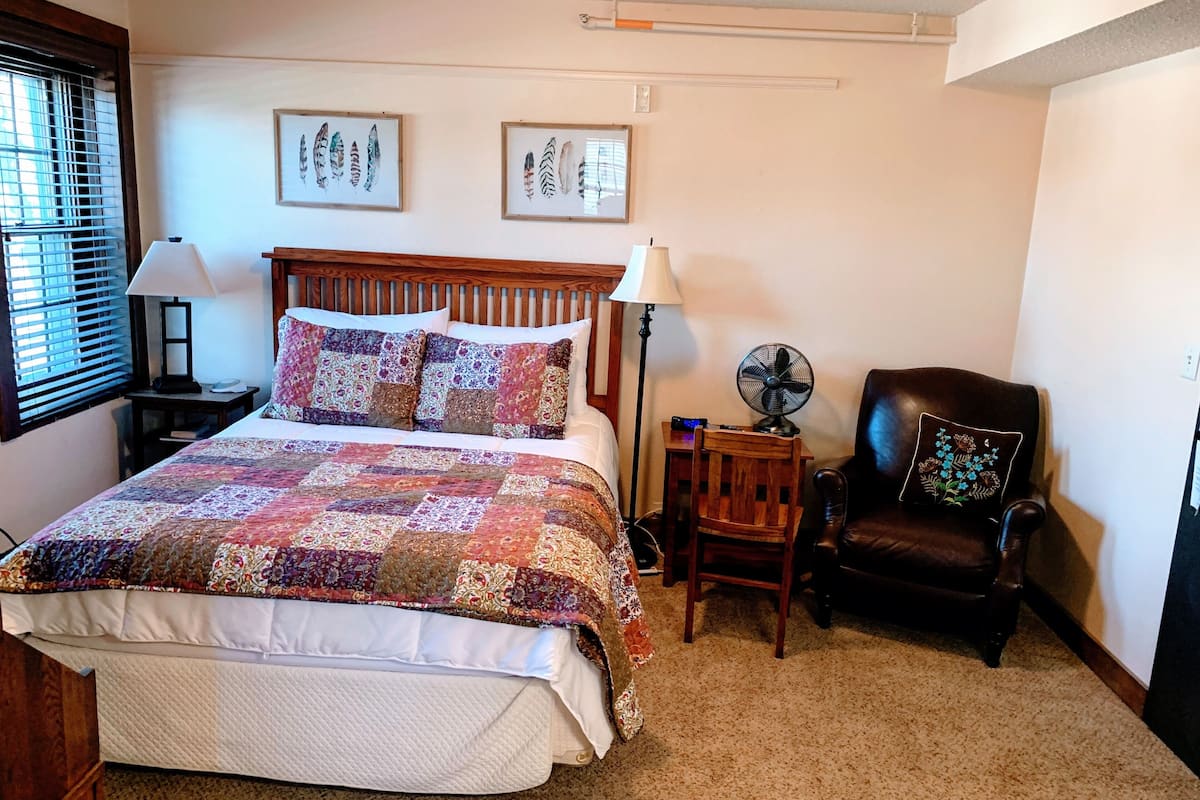 Un dormitorio con una cama, dos cuadros colgados en la pared, una silla y un ventilador de pie.