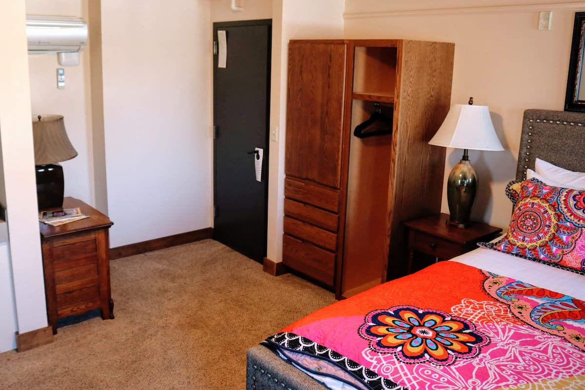 Habitación de hotel con cama, armario de madera, mesita de noche con lámpara y baño visible a través de una puerta abierta.