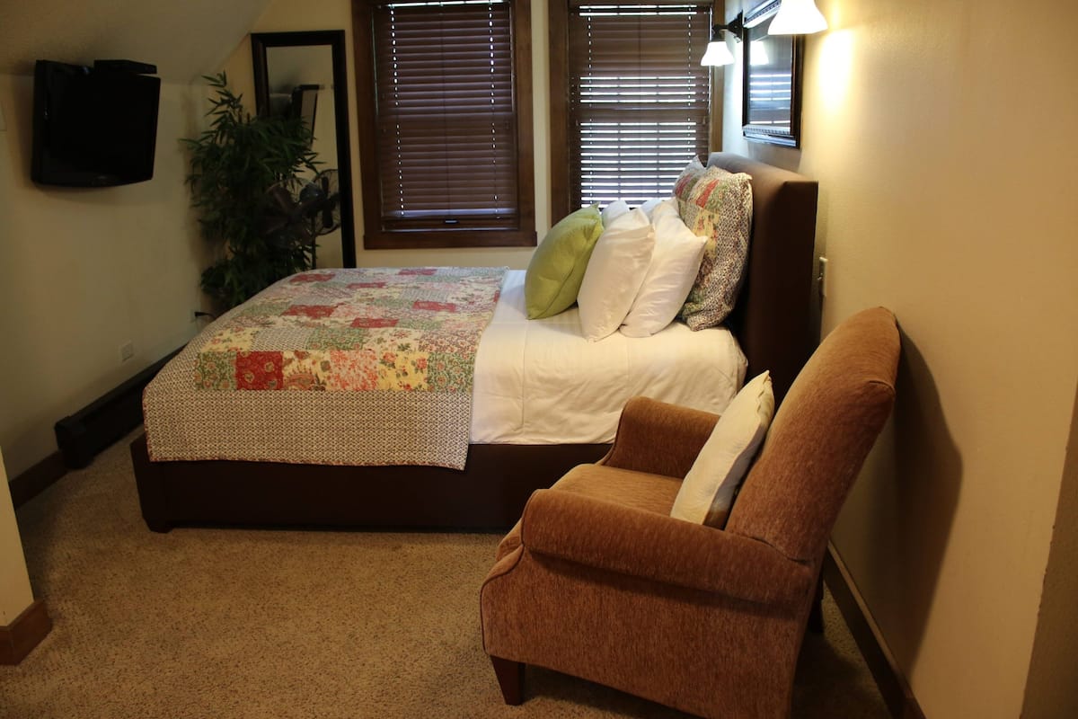 Un dormitorio con cama, una silla, un televisor y una ventana con persianas.
