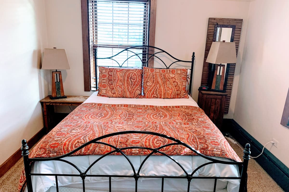 Una cama bien hecha con una colcha estampada y almohadas a juego, una mesita de noche con una lámpara, una ventana con persianas y un espejo en la pared.
