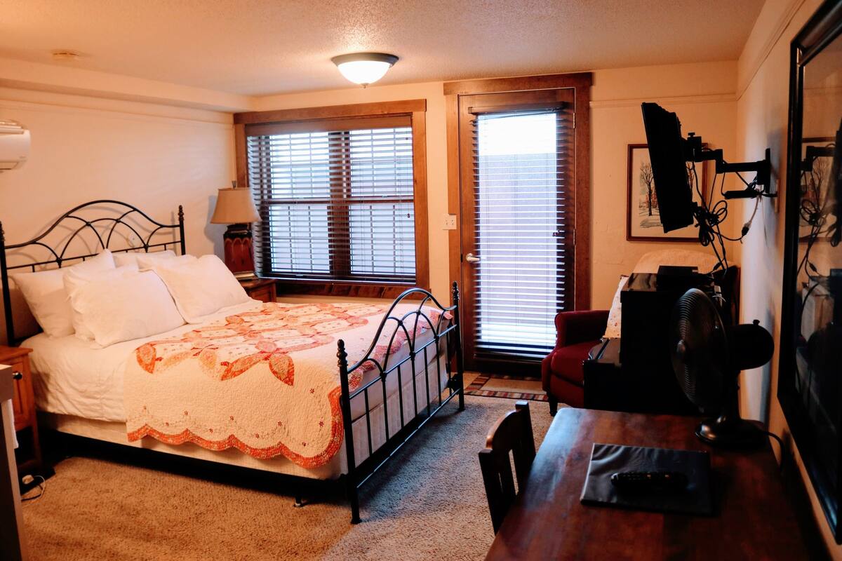 Un dormitorio con cama, mesita de noche, lámpara y escritorio con silla. Hay dos ventanas con persianas, una puerta y un televisor montado en la pared.