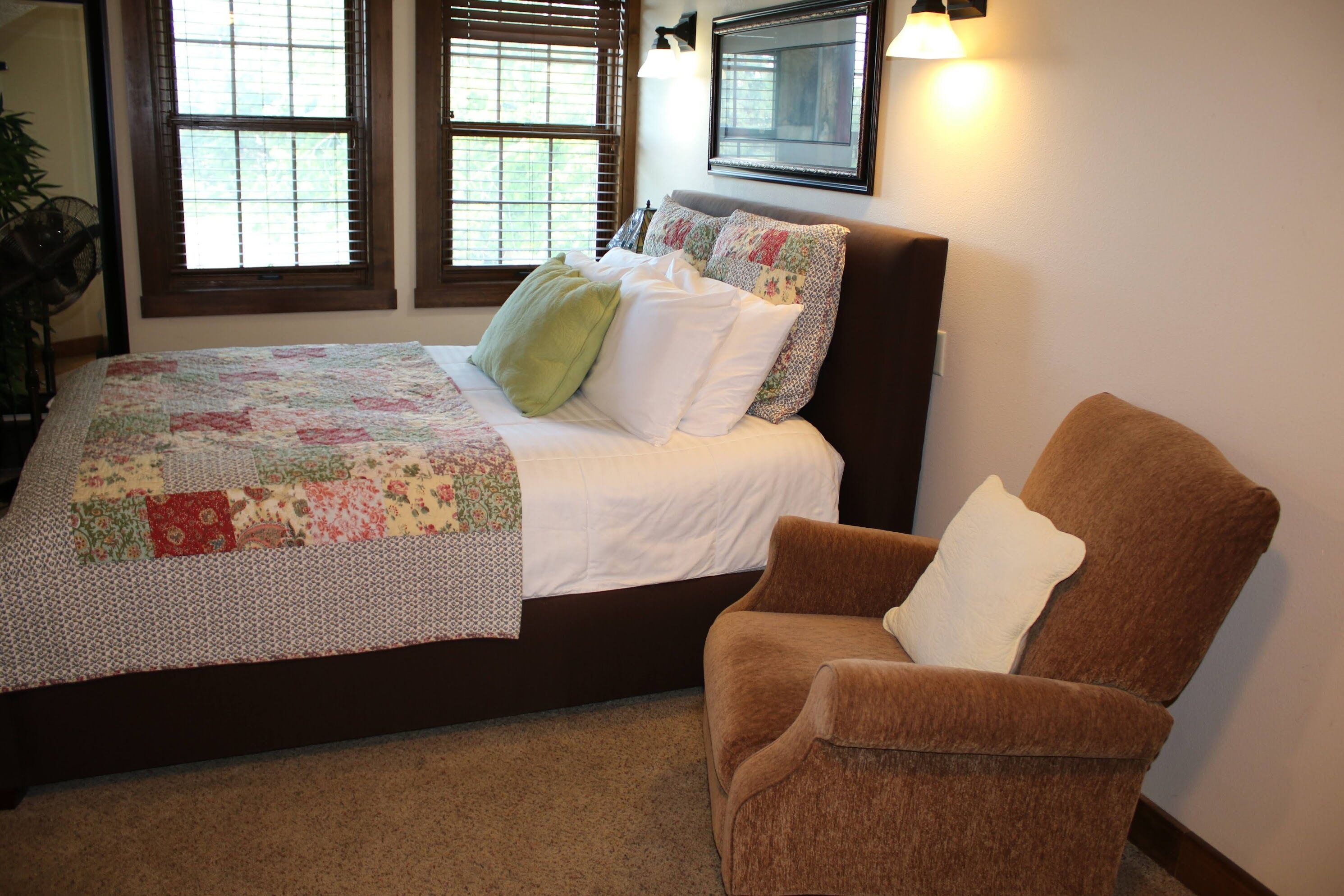 A bedroom with a bed, a chair, and two windows with blinds.