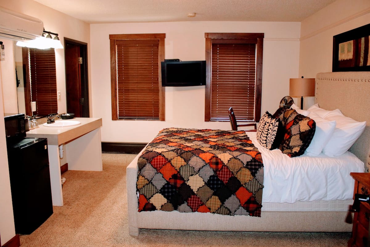 Un dormitorio con cama, mesita de noche, lámpara, televisión y baño con lavamanos y espejo.