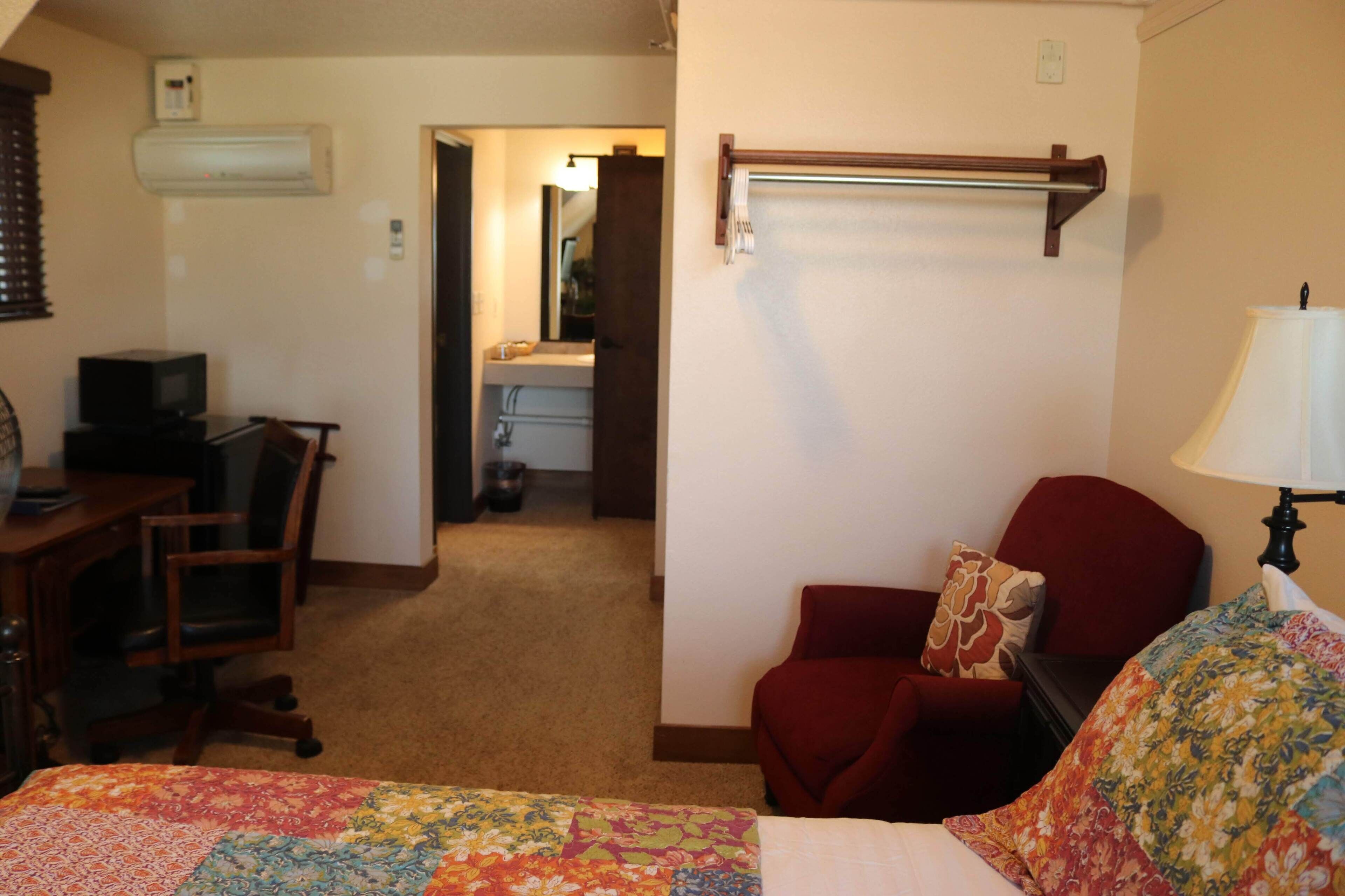 Une chambre d’hôtel avec un lit, un bureau avec un ordinateur, une chaise, un fauteuil rouge, une lampe et une salle de bain visible  à travers une porte ouverte.