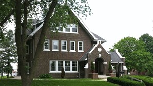 A two-story brick house with a gabled roof, a front entrance with a door, and a small porch with potted plants. - Iowa House (Ames)