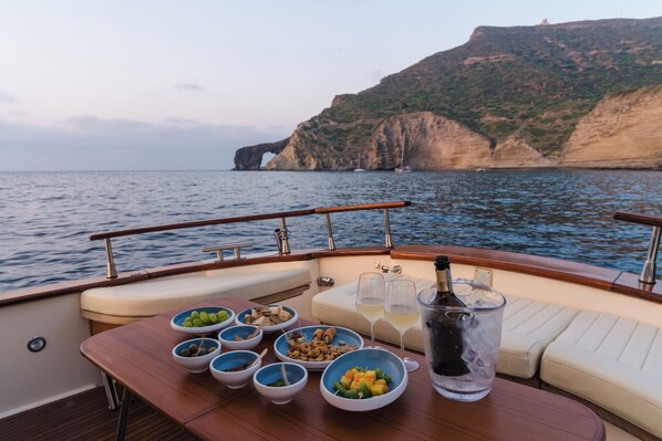 A boat with a dining table set with food and drinks, a bottle of champagne, and a scenic view of cliffs and water.