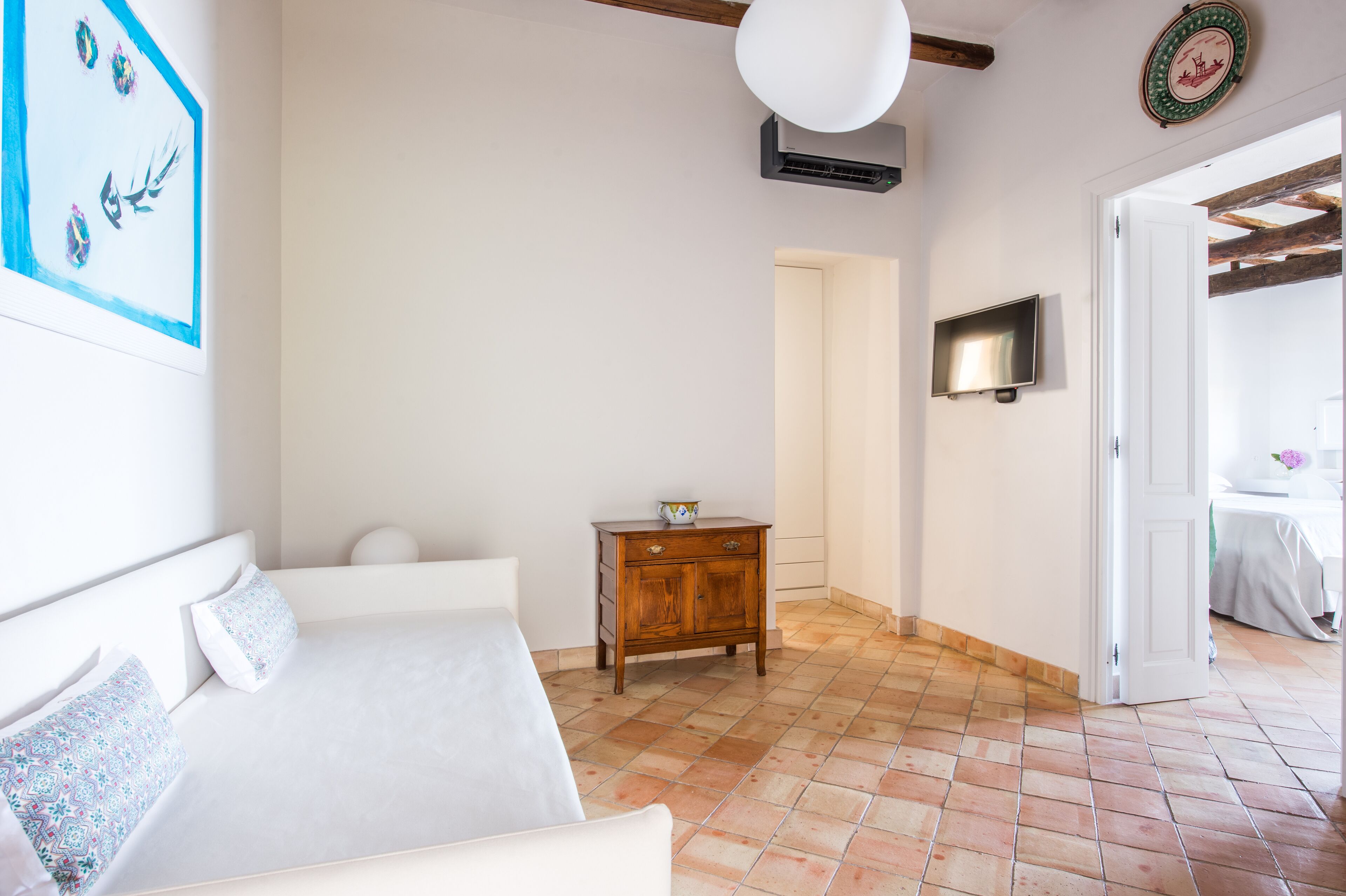 A bedroom with a bed, a wooden nightstand, a wall-mounted TV, and a door leading to another room.