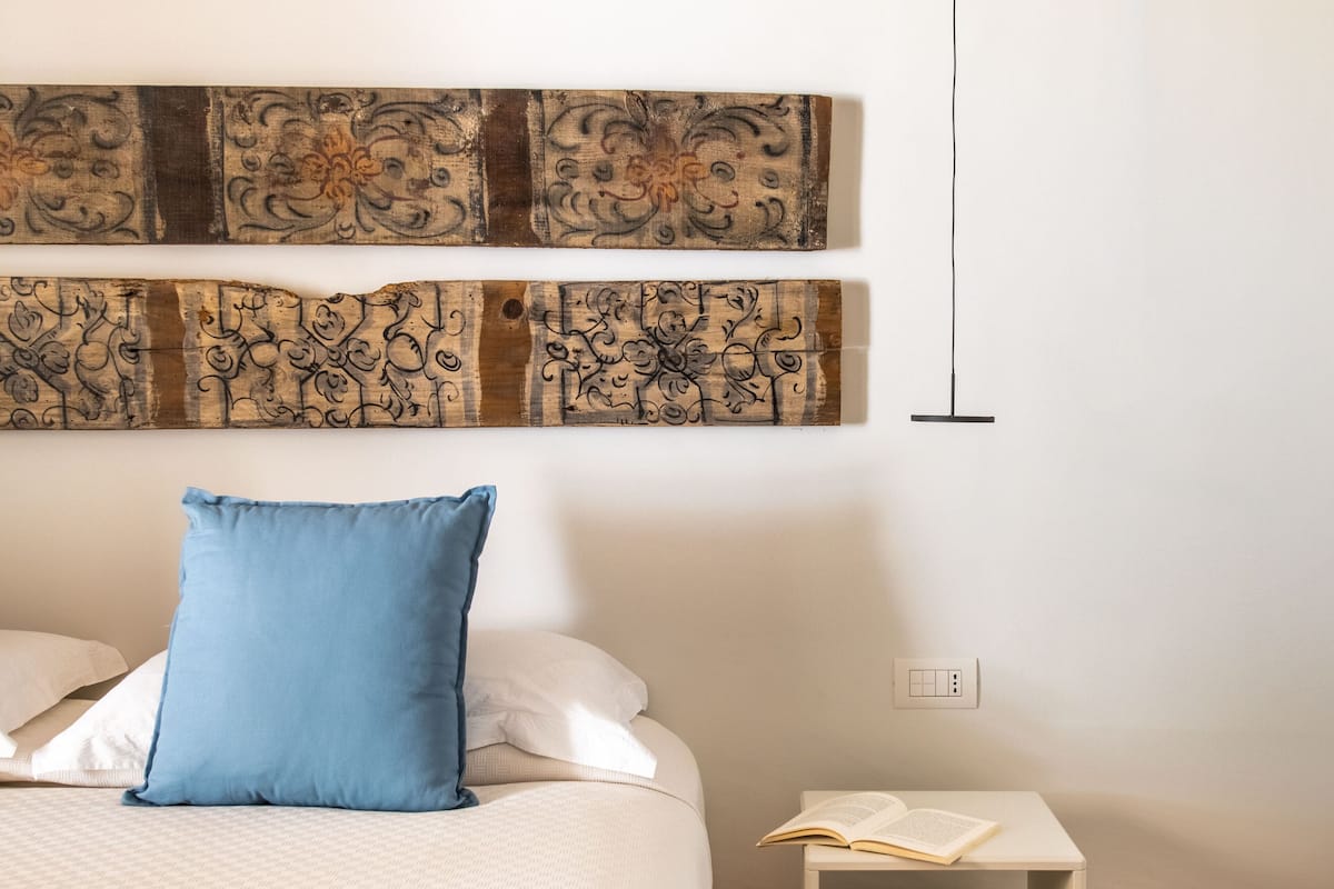 Une chambre à coucher avec un lit, un oreiller bleu, une table de chevet sur laquelle est posé un livre ouvert, et un mur orné d’œuvres d’art abstraites.