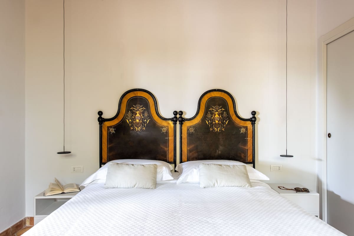 Un lit double avec une tête de lit en bois foncé, du linge de lit blanc et une table de chevet sur laquelle se trouvent une lampe et un livre.