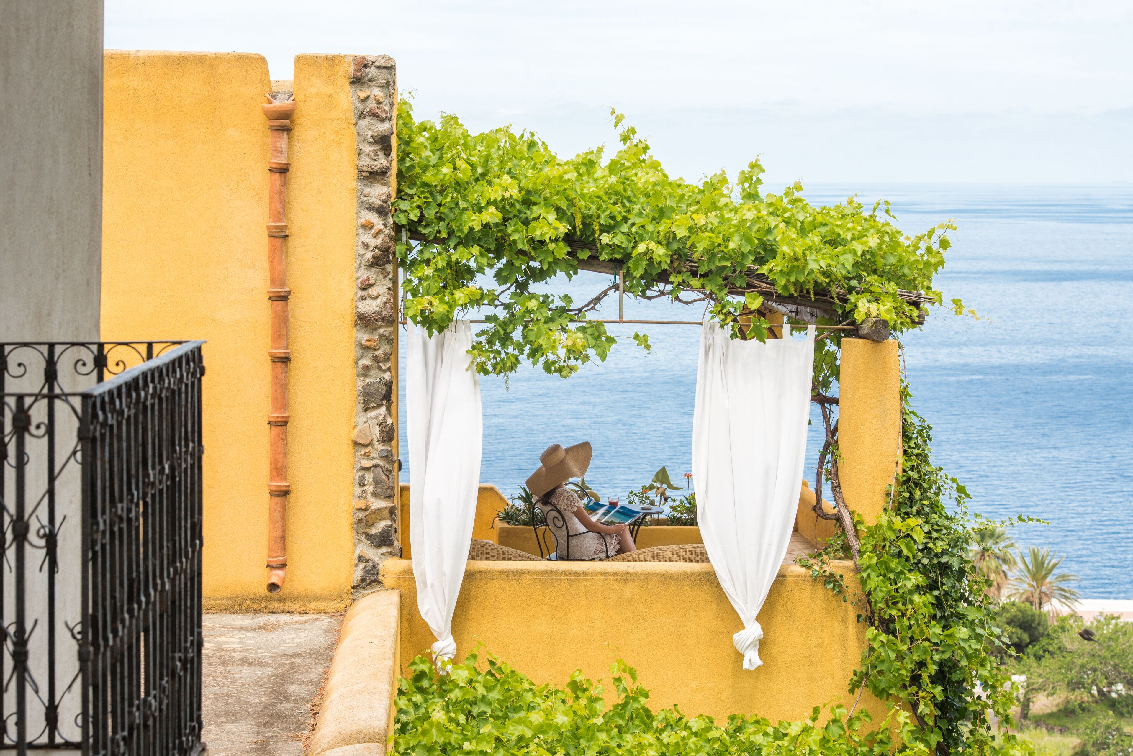 Eine Person sitzt an einem Tisch unter einer mit Weinreben bewachsenen Pergola mit Blick auf das Meer.