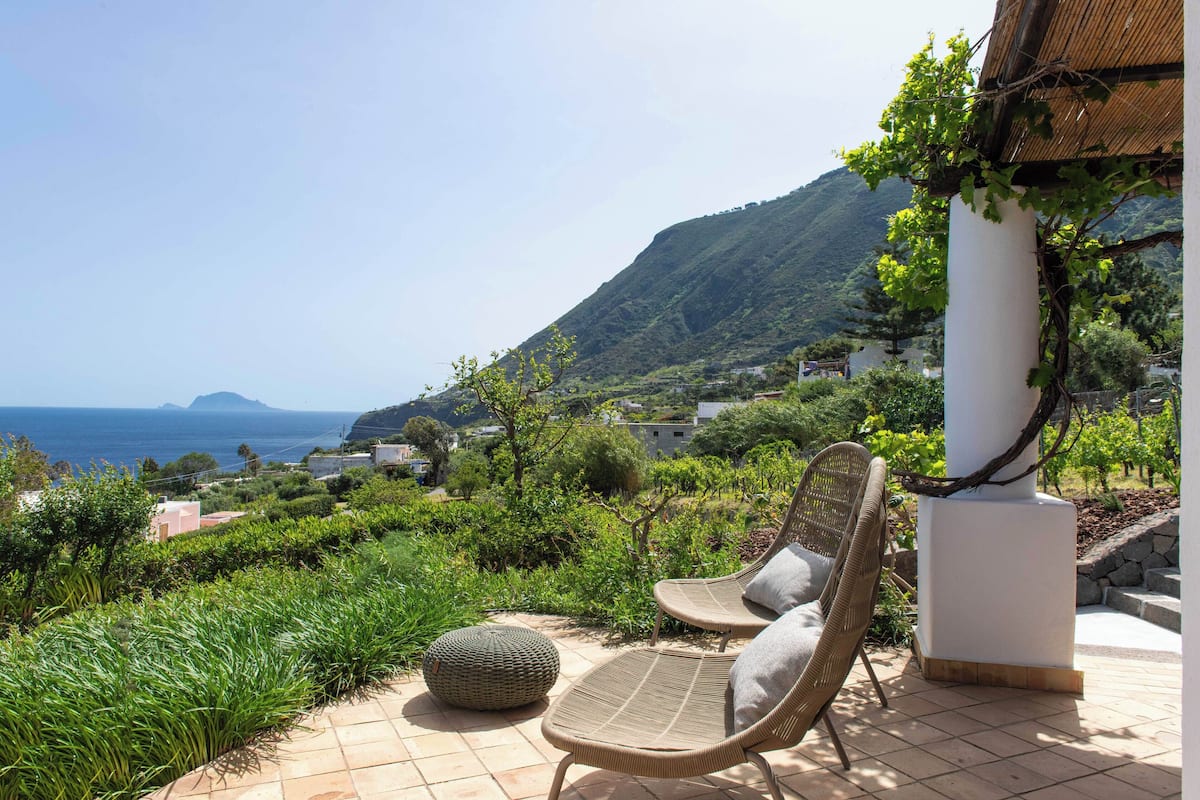 Une terrasse dotée de chaises en osier et d’un pouf rond, offrant une vue sur un vignoble et un paysage montagneux, avec la mer à l’horizon.