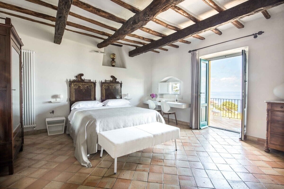 Une chambre à coucher avec un lit, un banc, un bureau et une vue sur l’extérieur par une porte ouverte.