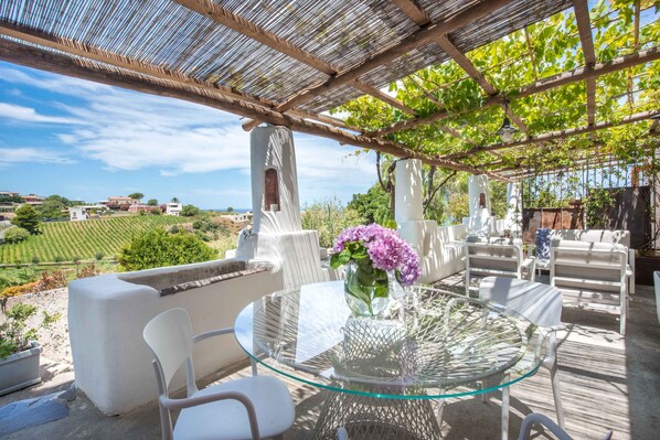 An outdoor patio with a glass table and white chairs, surrounded by a pergola with vines.