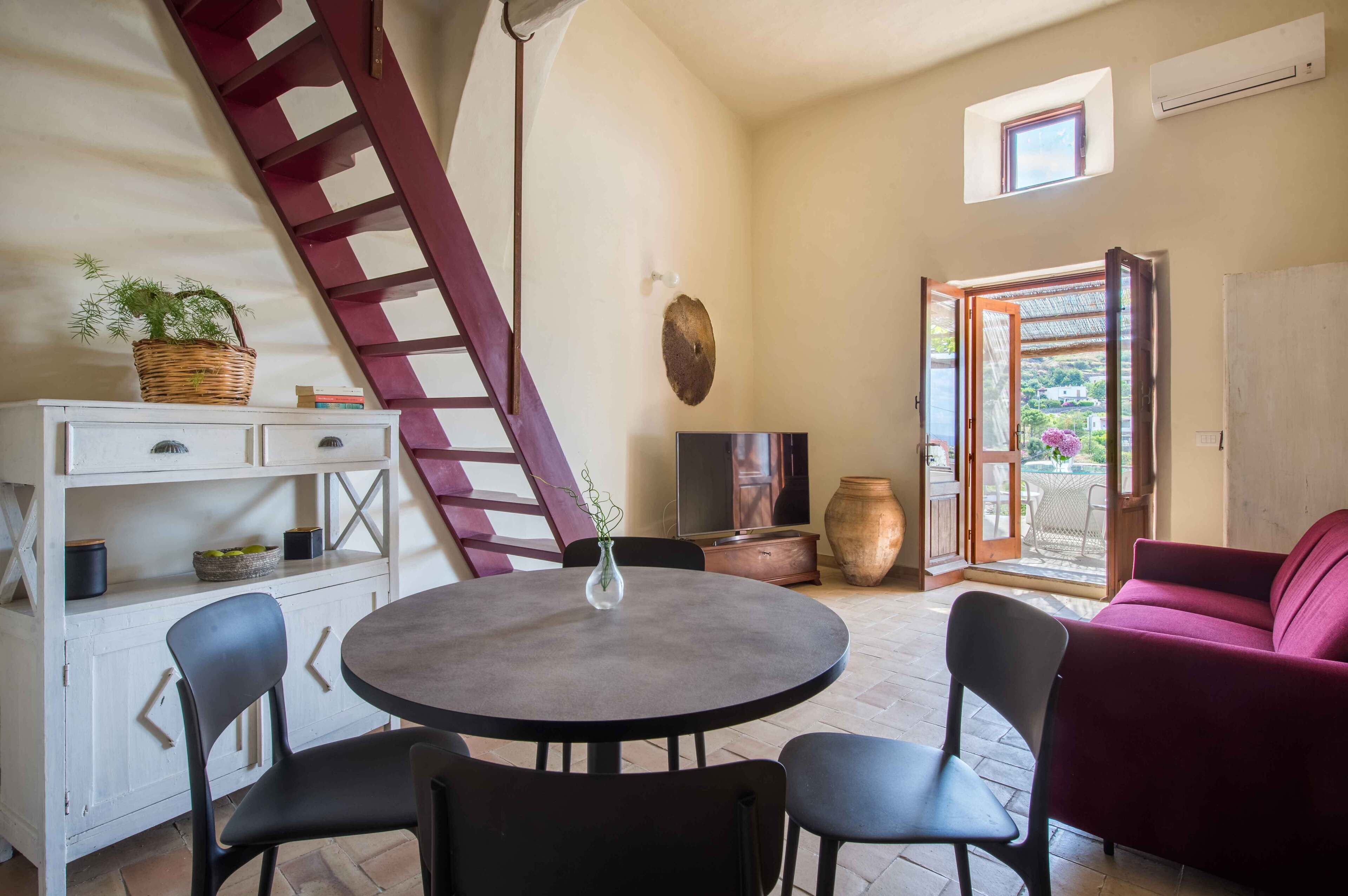 A cozy room with a round table, black chairs, a red sofa, a white cabinet, a wooden ladder, and a large window with a view of greenery.