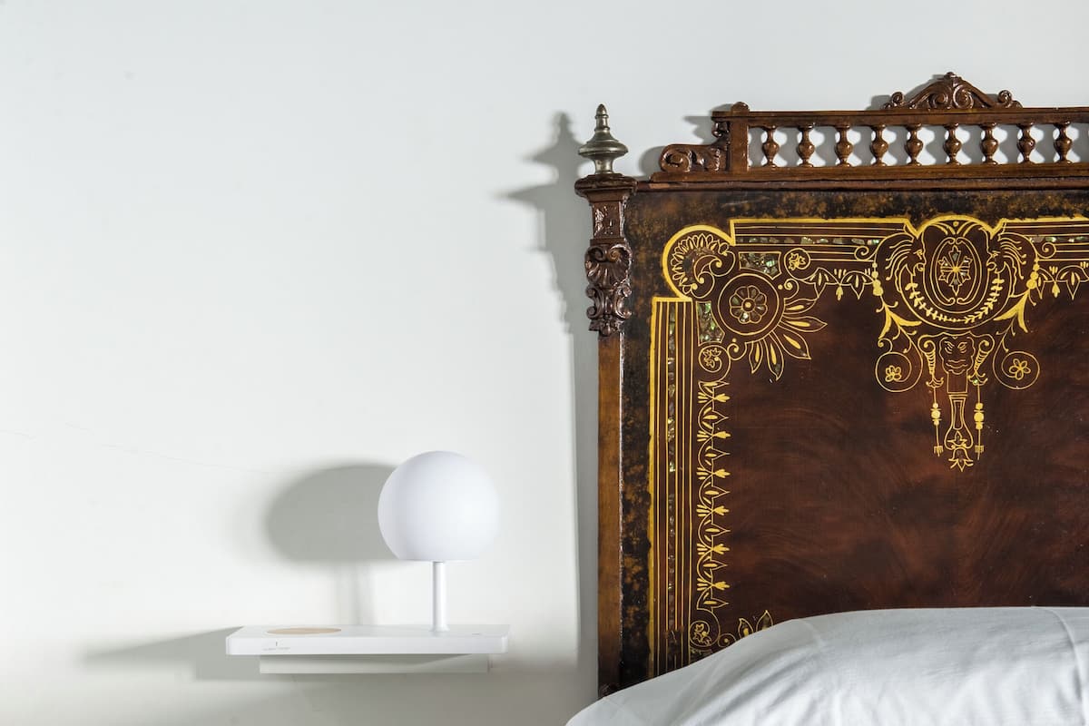Un lit bien fait, avec du linge de lit blanc, une tête de lit en bois ornée de motifs complexes, une table de chevet avec un livre, et une étagère murale sur laquelle repose un objet sphérique.