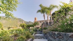 Una escalera de piedra conduce a un edificio con una torre de campanas, rodeado de abundante vegetación y plantas en flor.