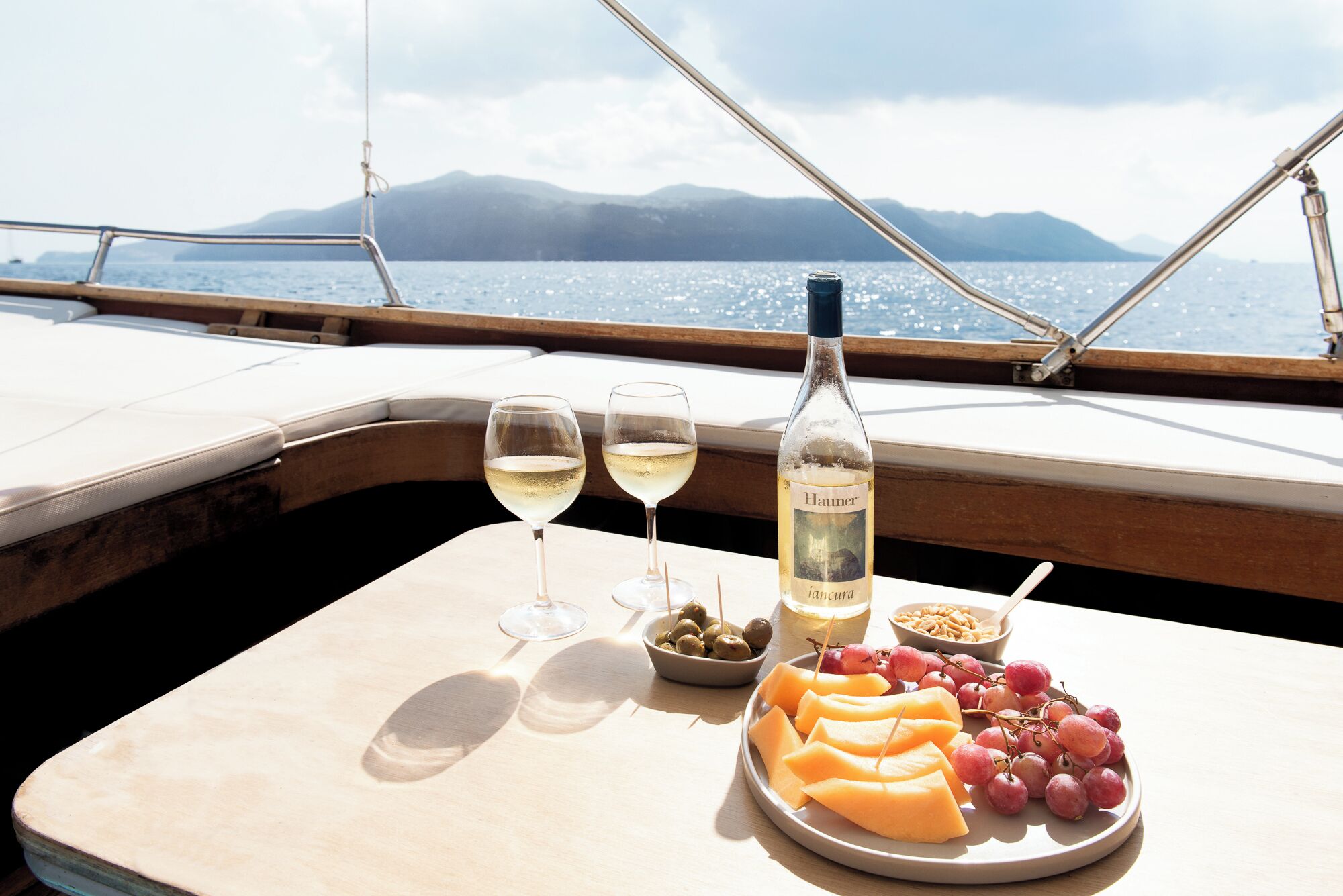 A table with a bottle of wine, two glasses, and a plate of cheese and grapes on a boat deck with a view of the sea and mountains.