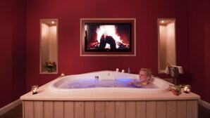 A room with a large bathtub, a television displaying a fireplace, and two wall-mounted sconces.