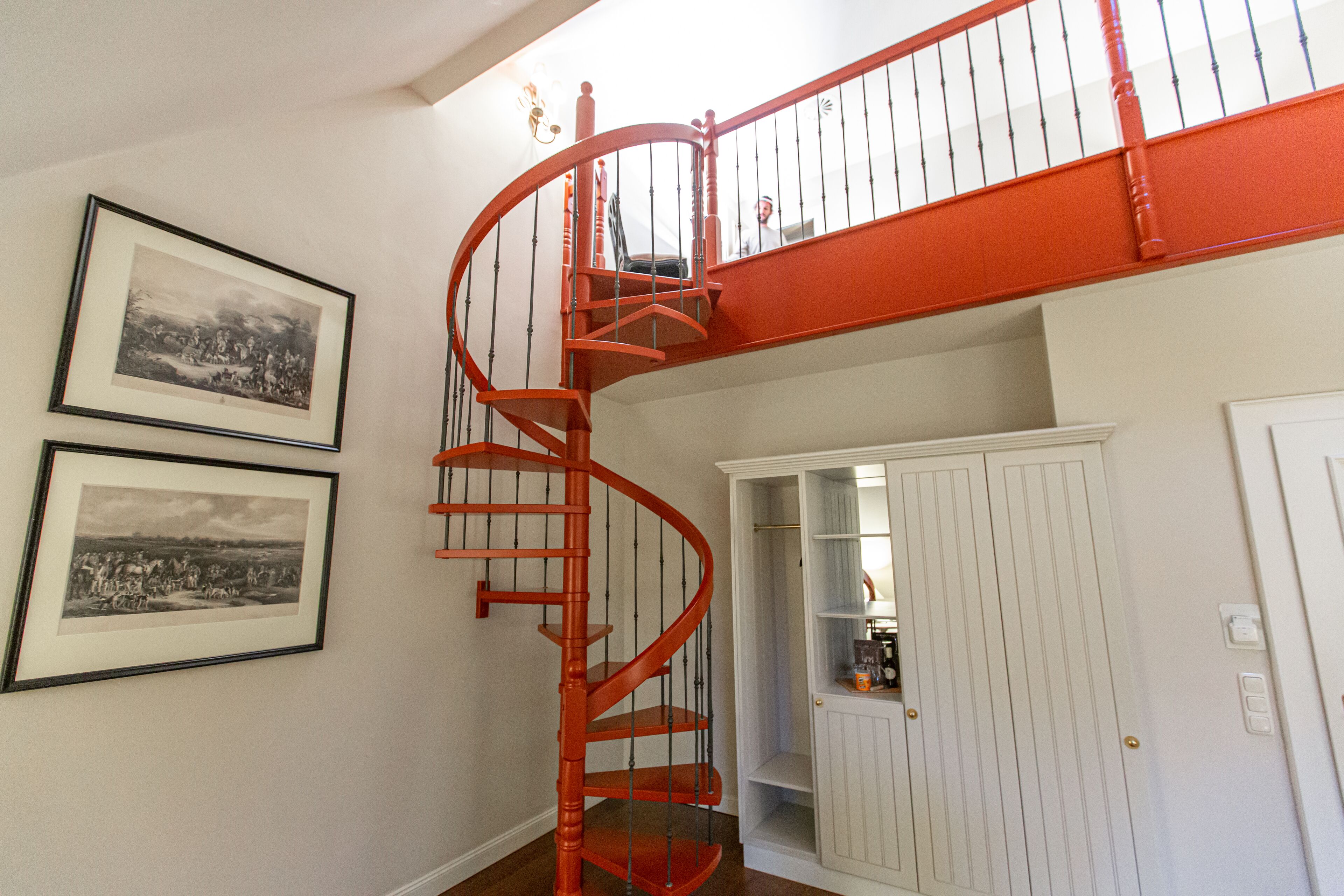 Eine Wendeltreppe mit roter Handlauf führt zu einer oberen Etage. Darunter befinden sich zwei gerahmte Schwarz-Weiß-Fotos an der Wand.