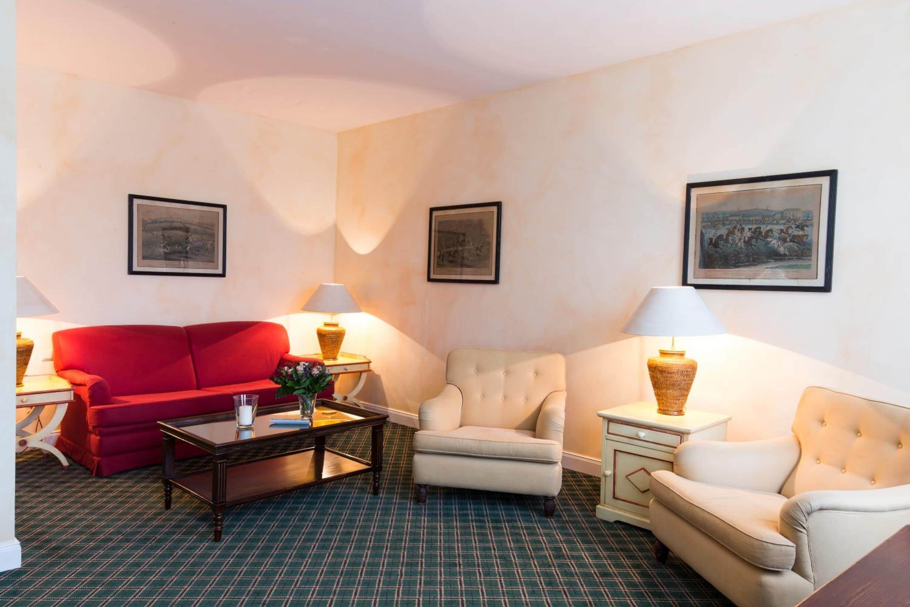 Une chambre d’hôtel avec un canapé rouge, un fauteuil beige, une petite table et deux lampes.