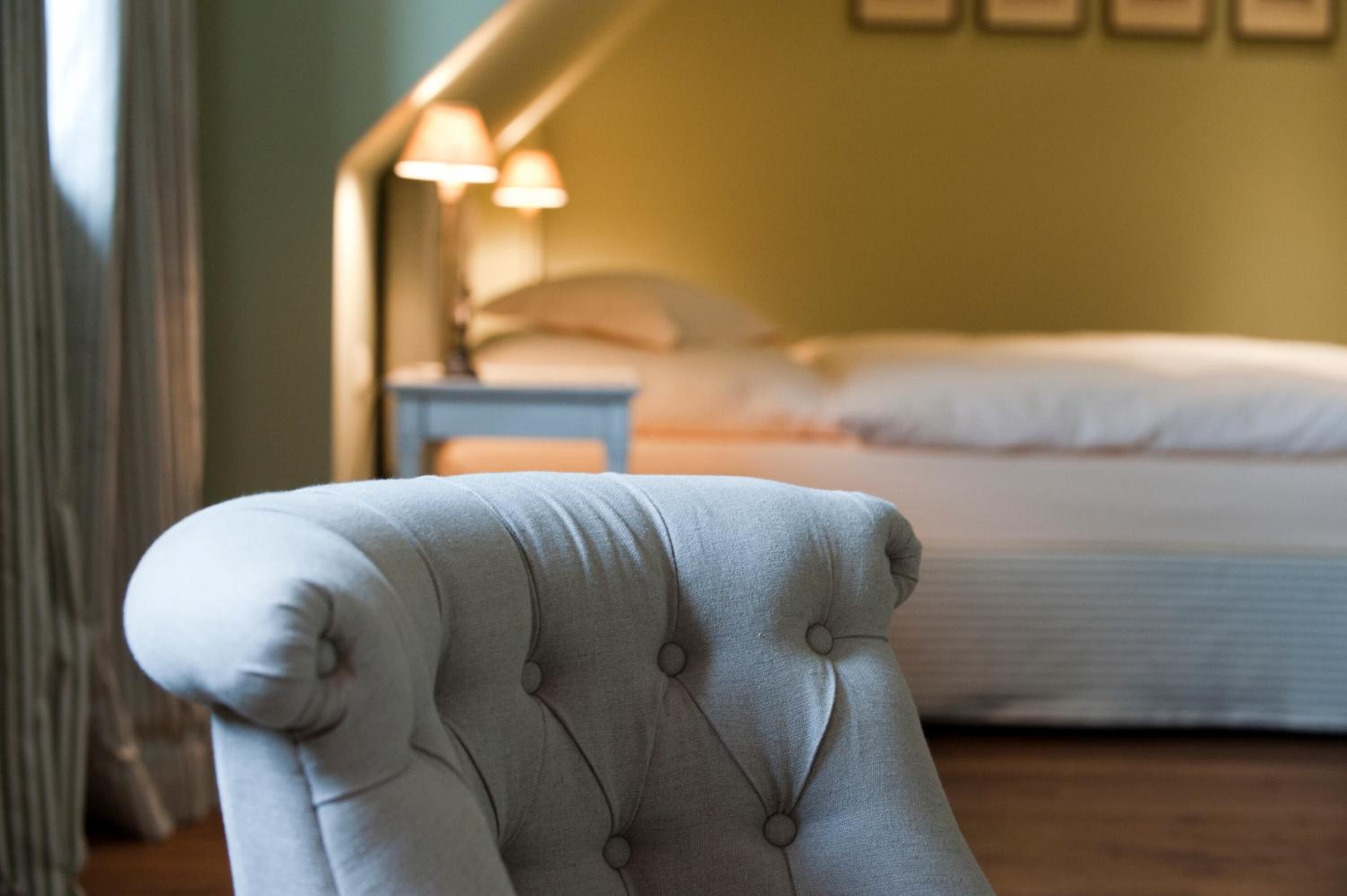 A tufted armchair in a room with a bed, a nightstand, and wall-mounted lamps.
