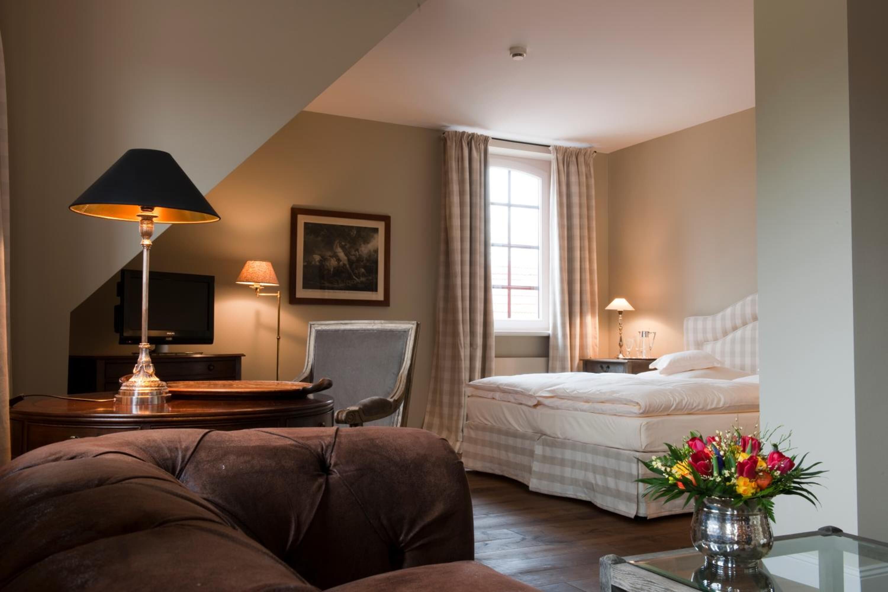 A hotel room with a bed, a sofa, a desk with a lamp, a chair, a TV, and a window with curtains.