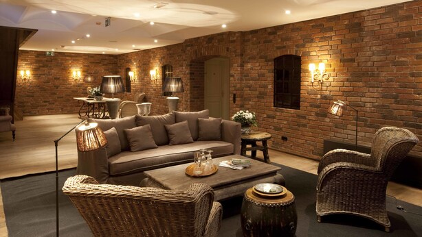 A cozy lounge area with a brick wall, a sofa, a coffee table, wicker chairs, and ambient lighting.