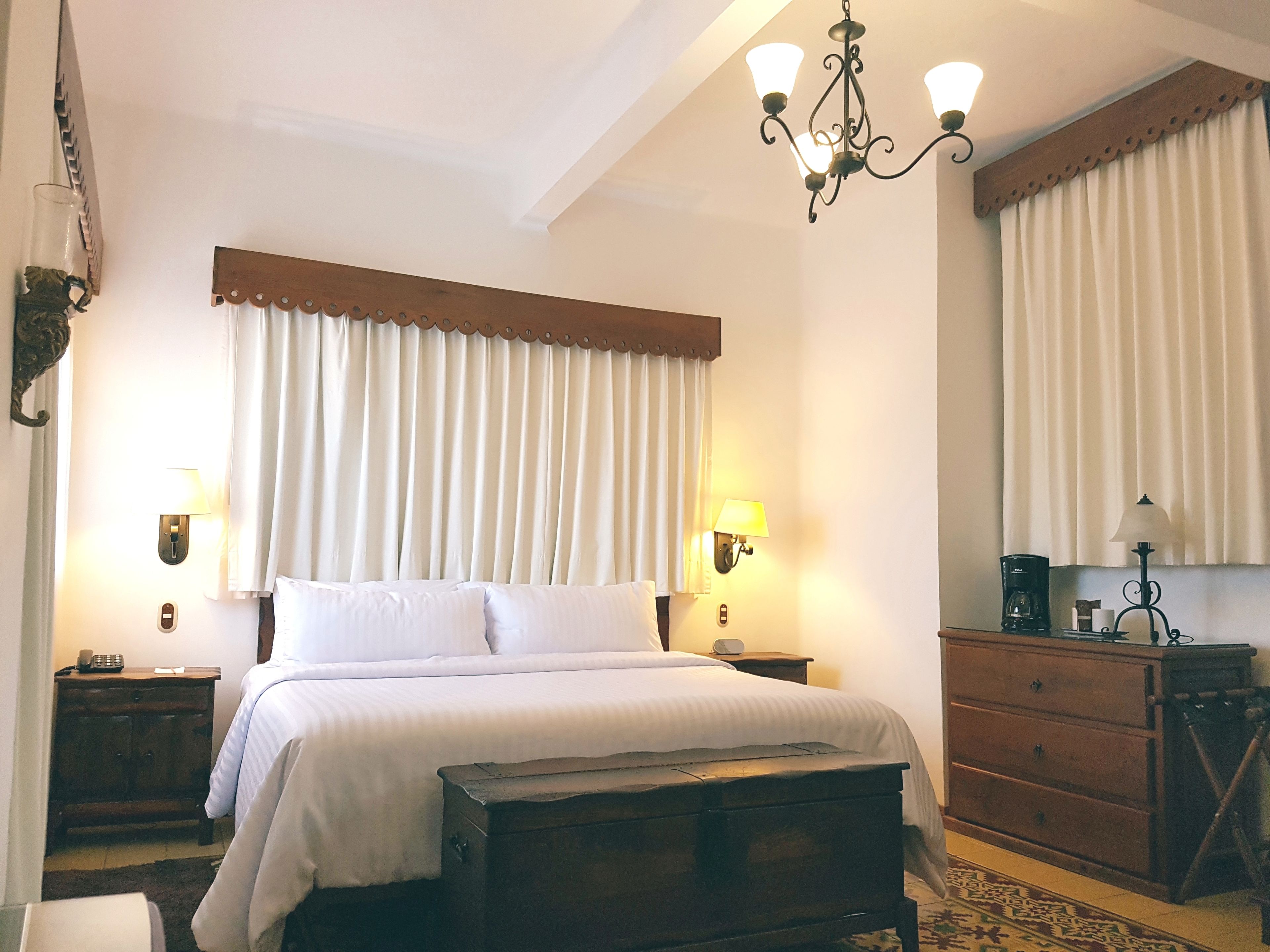 Una habitación de hotel con una cama grande, dos mesitas de noche con lámparas, un aparador de madera y un candelabro.