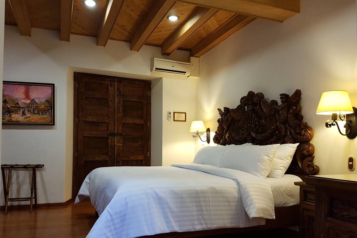 A bedroom with a wooden bed, a large wooden door, an air conditioning unit, a wall-mounted lamp, and a framed picture on the wall.
