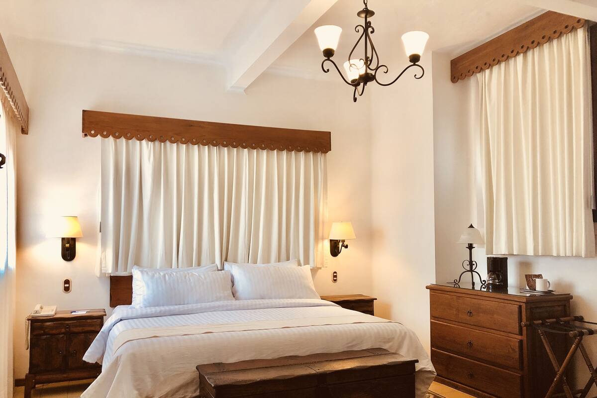 A hotel room with a bed, bedside lamps, a wooden dresser, a mirror, and a chandelier.
