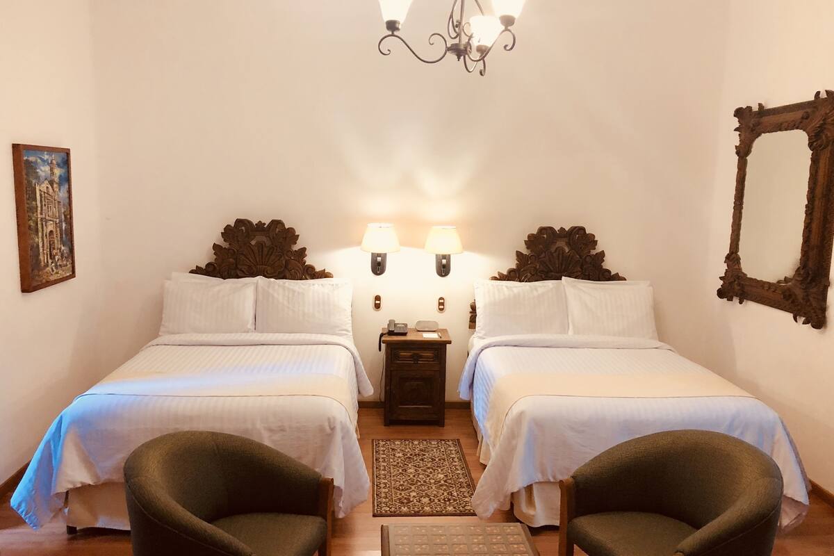 A room with two beds, a small table, two chairs, a nightstand with lamps, and a chandelier.