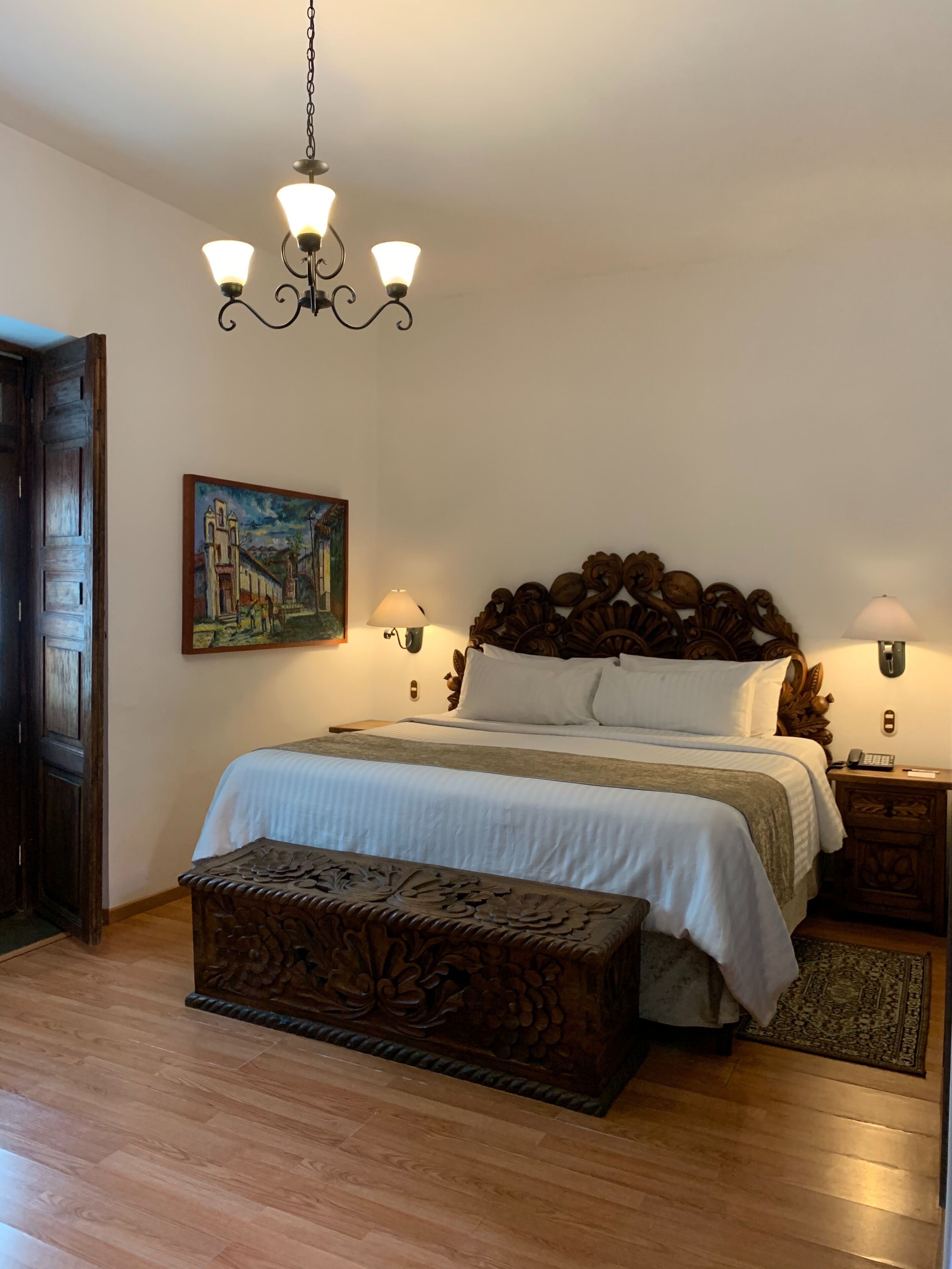 Un dormitorio con una cama de madera, un banco, un cuadro y una araña.