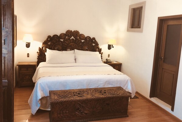 Quarto com uma grande cabeceira de madeira, duas mesinhas de cabeceira com luminárias, um banco com entalhes e uma porta.