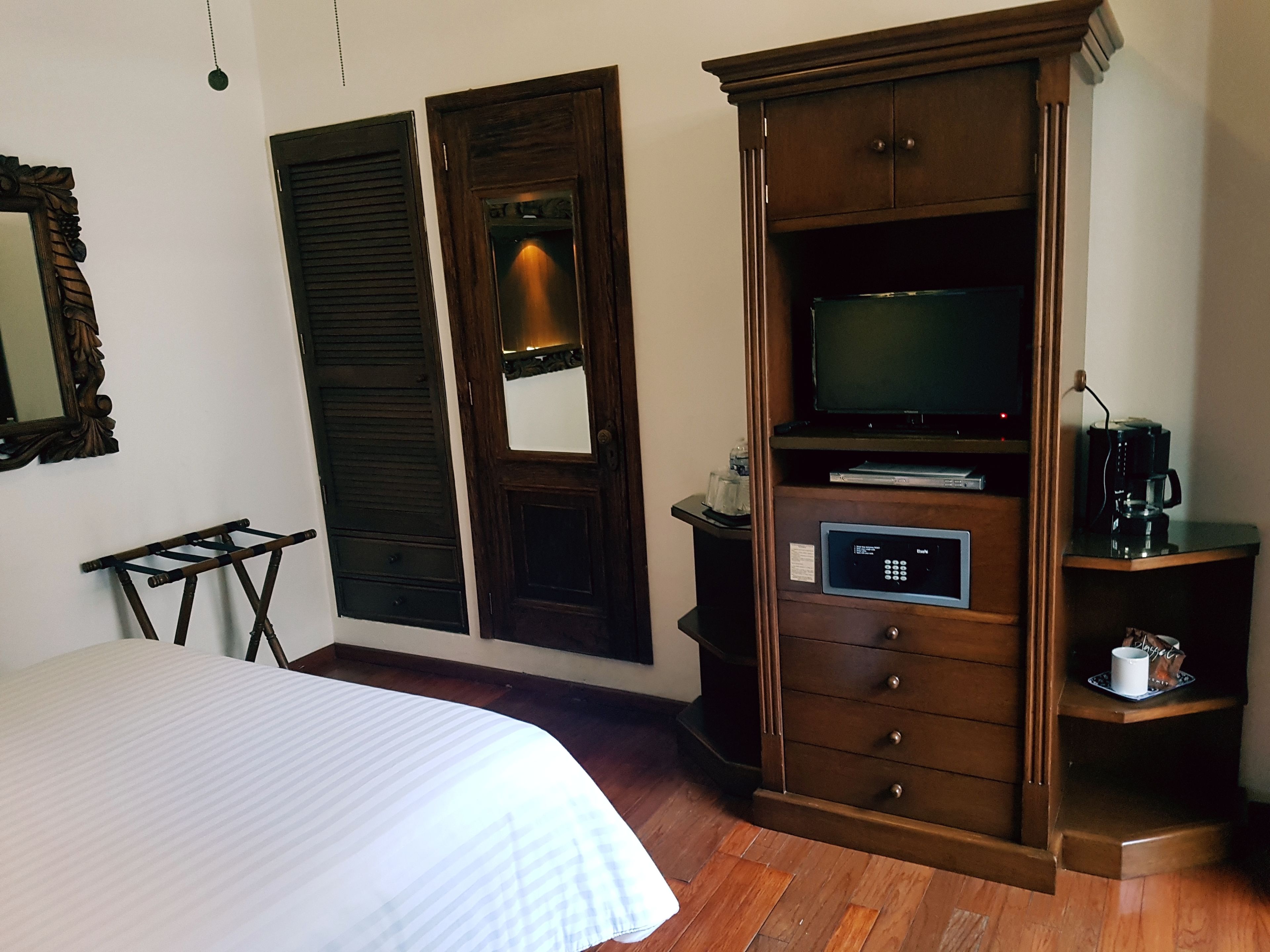 A hotel room with a bed, a wooden wardrobe with a television and a coffee machine, and a small table with a bottle.