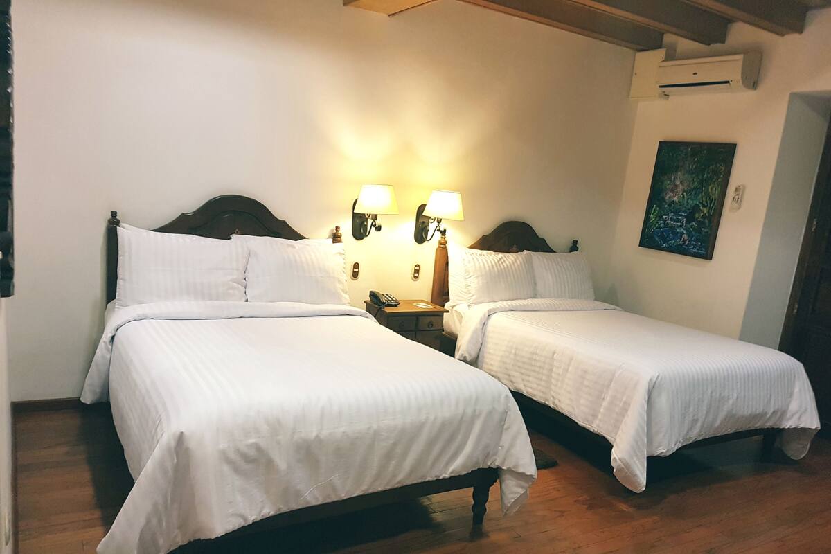 A double room with two beds, wooden floors, and a nightstand with a lamp between them.