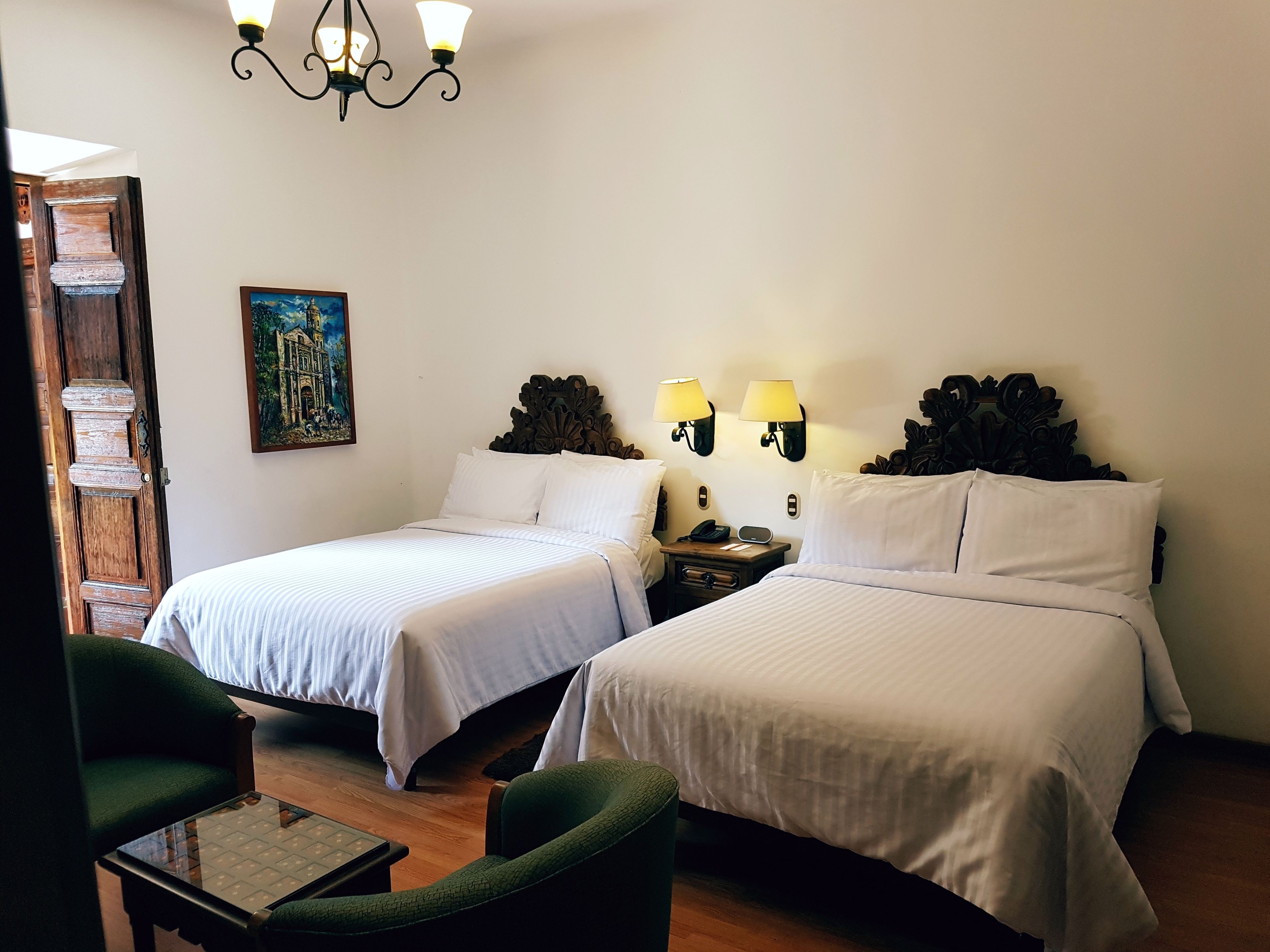 A hotel room with two single beds, a small table, two chairs, a nightstand with a lamp, and a painting on the wall.