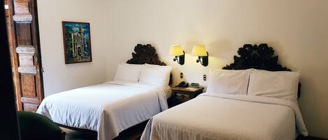 A hotel room with two single beds, a small table, two chairs, a nightstand with a lamp, and a painting on the wall.