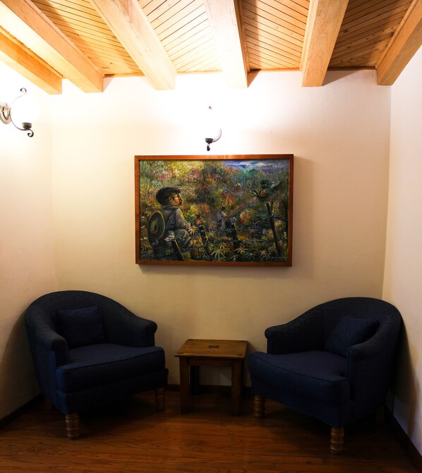 Une pièce avec des poutres en bois au plafond, deux fauteuils bleu foncé, une petite table en bois et un tableau coloré accroché au mur.