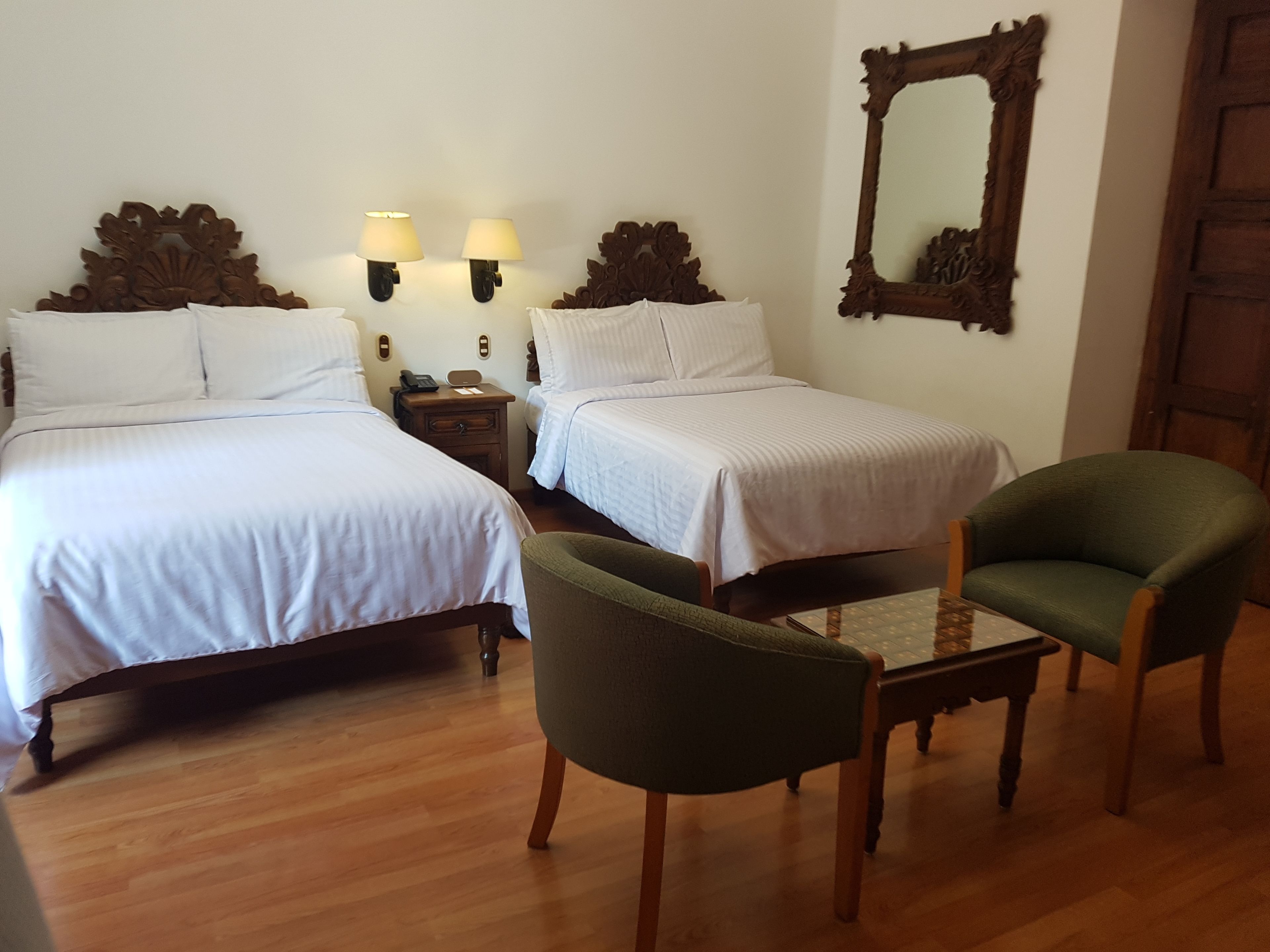 Una habitación de hotel con dos camas individuales, un sillón verde, una mesa de madera y un espejo en la pared.